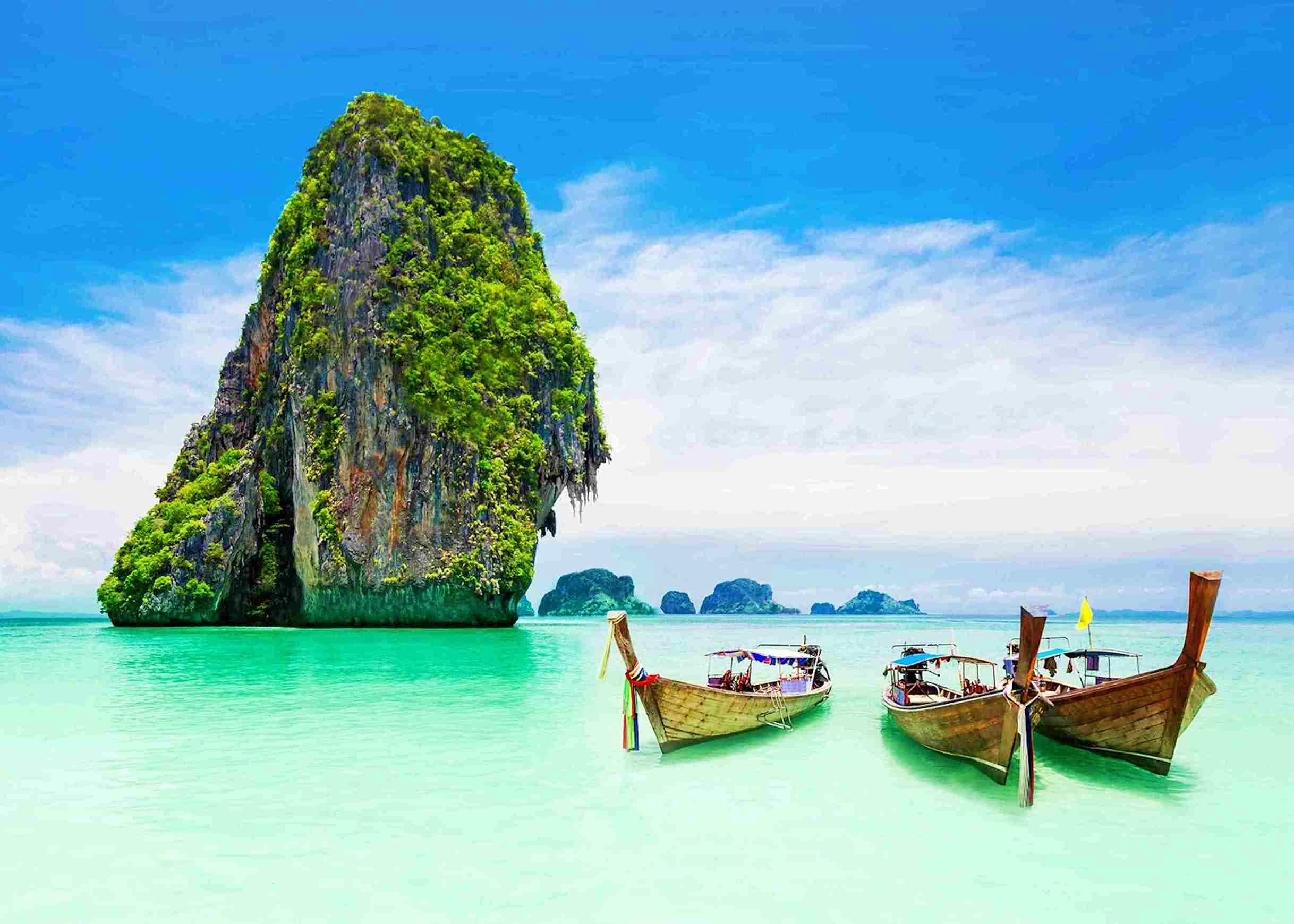 Limestone cliff covered in green vegetation rising from turquoise water with traditional long tail boats in shallow tropical 